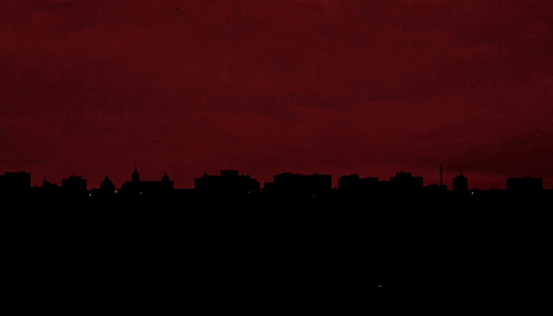 Dark brick-red duotone photograph showing the outlines of a city skyline at dusk in deep maroon and black tones